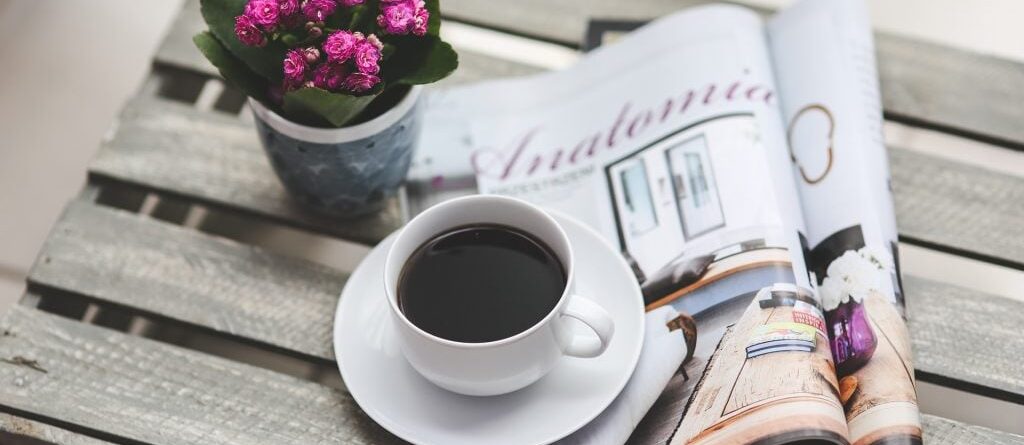 Es ist ein aufgeschlagenes Magazin auf einer Holzkiste zu sehen, teils auf dem Magazin steht eine gefüllte Kaffeetasse. Links daneben steht ein kleiner Blumentopf mit mit pinken Blüten.