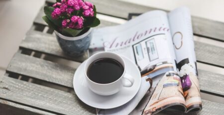 Es ist ein aufgeschlagenes Magazin auf einer Holzkiste zu sehen, teils auf dem Magazin steht eine gefüllte Kaffeetasse. Links daneben steht ein kleiner Blumentopf mit mit pinken Blüten.