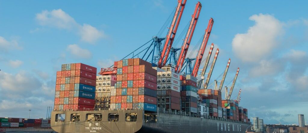 Es ist ein großes Containerschiff von hinten-schräg zu sehen. Es liegt in einem Containerhafen an.