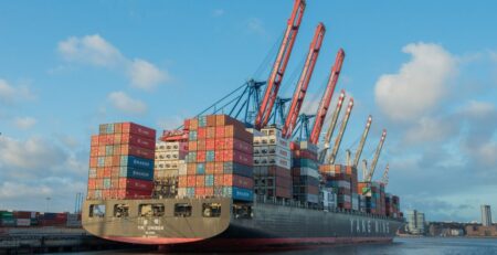 Es ist ein großes Containerschiff von hinten-schräg zu sehen. Es liegt in einem Containerhafen an.