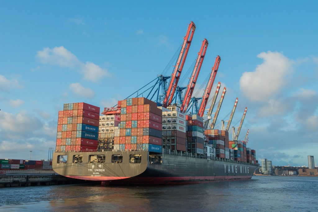 Es ist ein großes Containerschiff von hinten-schräg zu sehen. Es liegt in einem Containerhafen an.