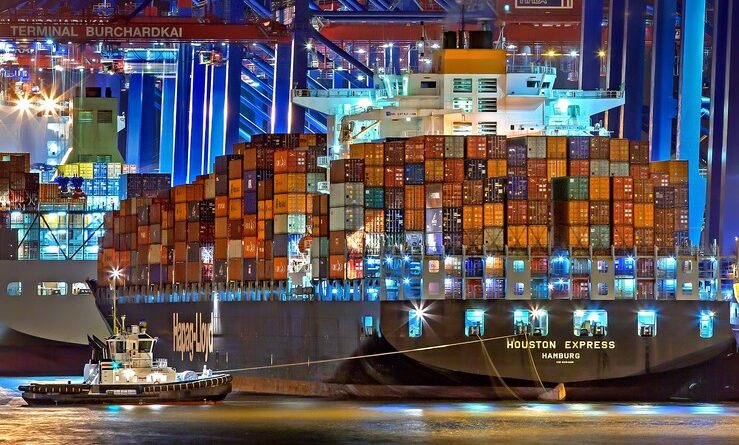 Es ist ein riesiges, voll beleuchtest Containerschiff zu sehen im Hafen angelegt. Auch ein Schlepper ist zu sehen.