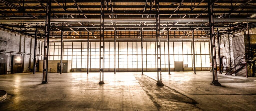 Es ist eine leere Lagerhalle zu sehen in der das Sonnenlicht leicht durch die verschmutzten Fenster scheint