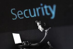 Es ist ein Mann in einem dunklen Raum im Profil zu sehen wie er an einem Laptop arbeitet. Über ihm steht im LED Style Security