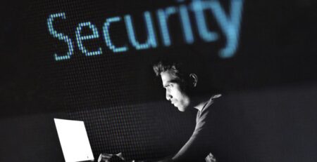 Es ist ein Mann in einem dunklen Raum im Profil zu sehen wie er an einem Laptop arbeitet. Über ihm steht im LED Style Security