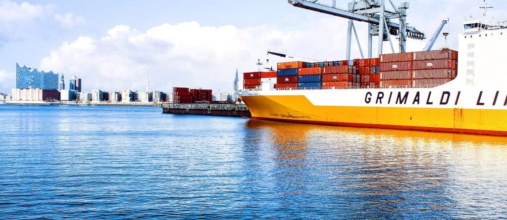 Es ist ein Containerschiff, liegend, in einem Containerhafen zu sehen. Im Hintergrund ist eine Stadt.