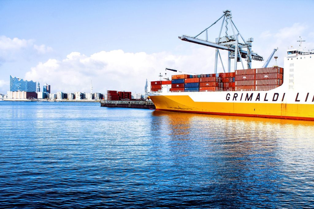 Es ist ein Containerschiff, liegend, in einem Containerhafen zu sehen. Im Hintergrund ist eine Stadt.