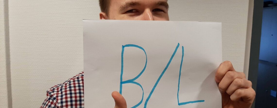 Junger Mann der den Daumen hoch macht und ein Blatt Papier vor sein "halbes" Gesicht hält auf dem in Groß "B/L" steht