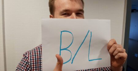 Junger Mann der den Daumen hoch macht und ein Blatt Papier vor sein "halbes" Gesicht hält auf dem in Groß "B/L" steht