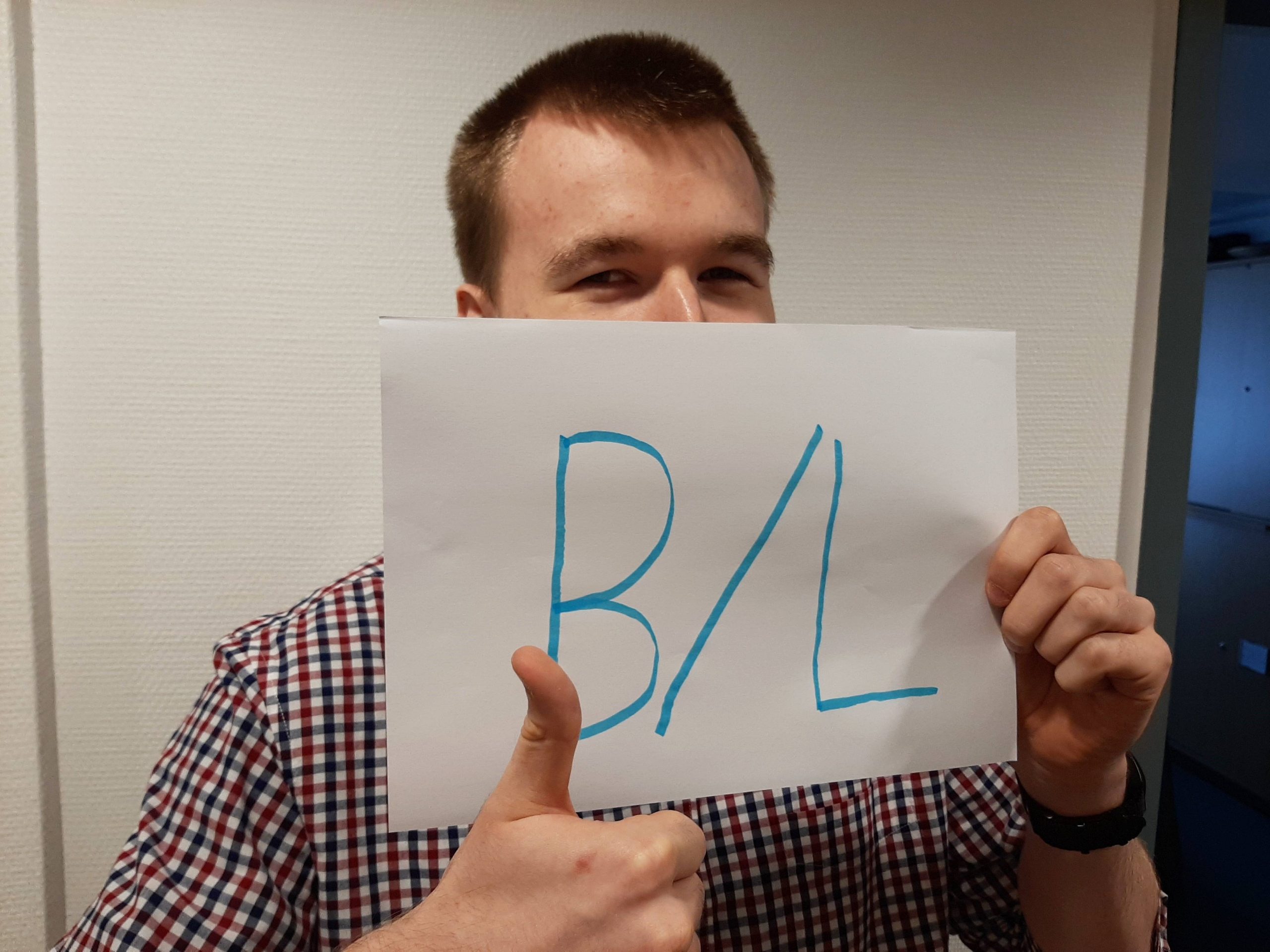 Junger Mann der den Daumen hoch macht und ein Blatt Papier vor sein "halbes" Gesicht hält auf dem in Groß "B/L" steht